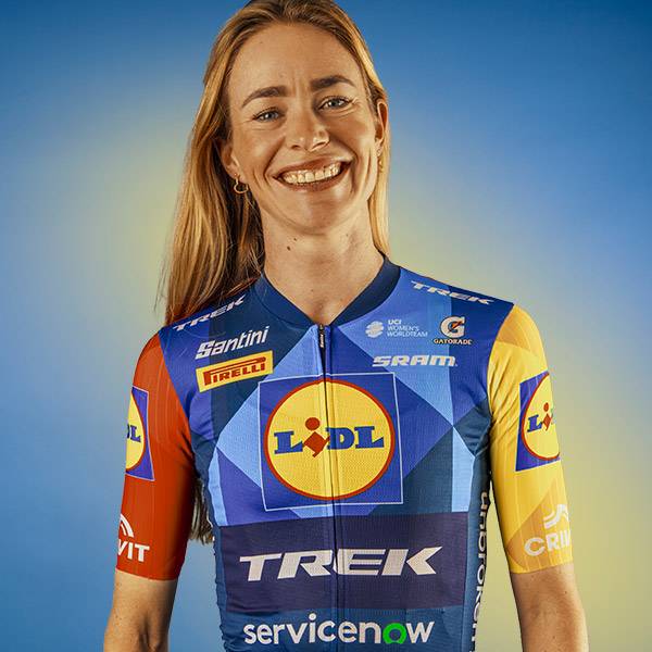 Vrouw in een blauw, geel en rood fietsshirt met geometrische patronen en sponsorlogo's.