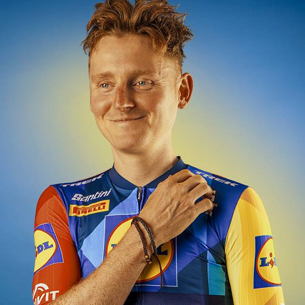 Een glimlachende man in een blauw-geel sportshirt met logo's en een gevlochten armband.