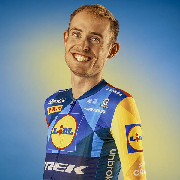 Lachende man in een blauw en geel fietsshirt met geometrische patronen en logo's.