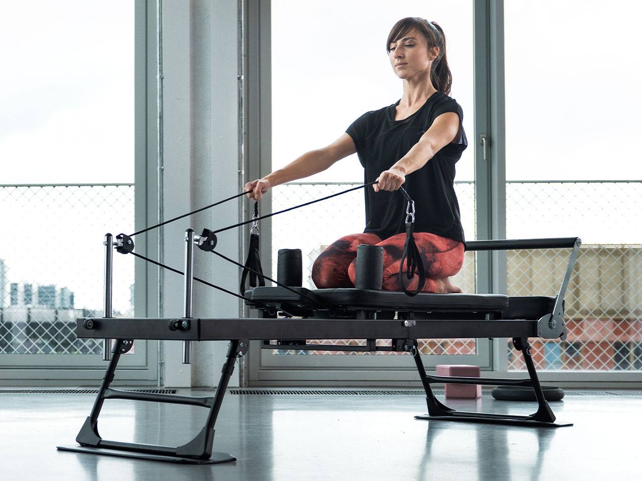Vrouw doet Pilates op een reformer met grote ramen op de achtergrond.