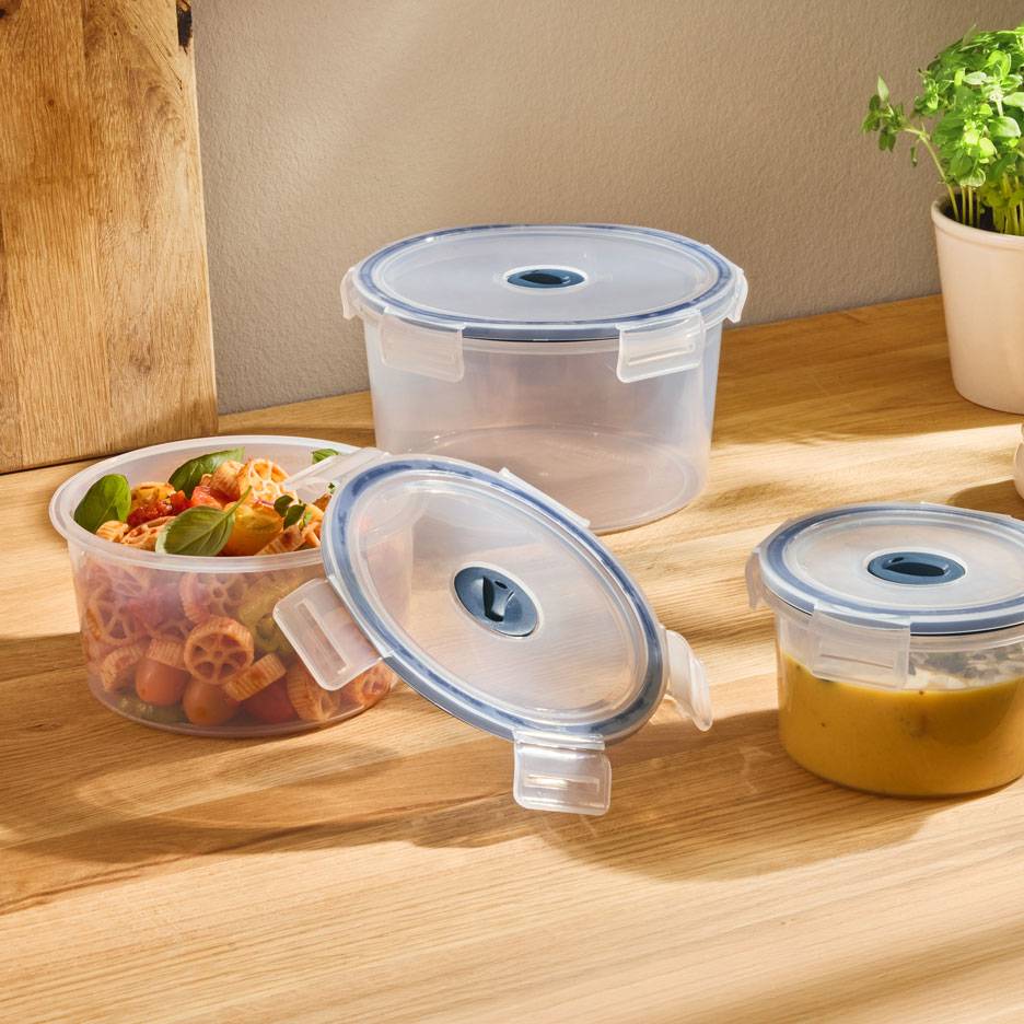 Doorzichtige ronde voedselcontainers met blauwgerande deksels, één gevuld met pastasalade, een andere met soep.