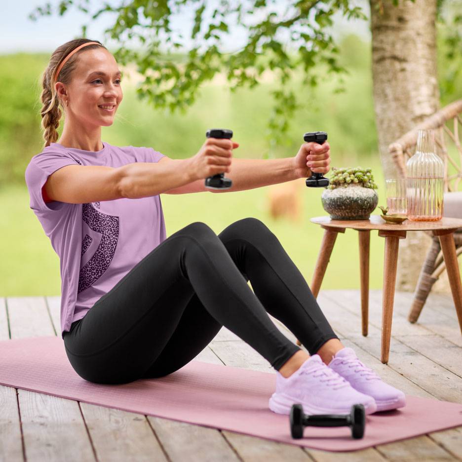 Vrouw in paars T-shirt en zwarte legging oefent met dumbbells op een yogamat.