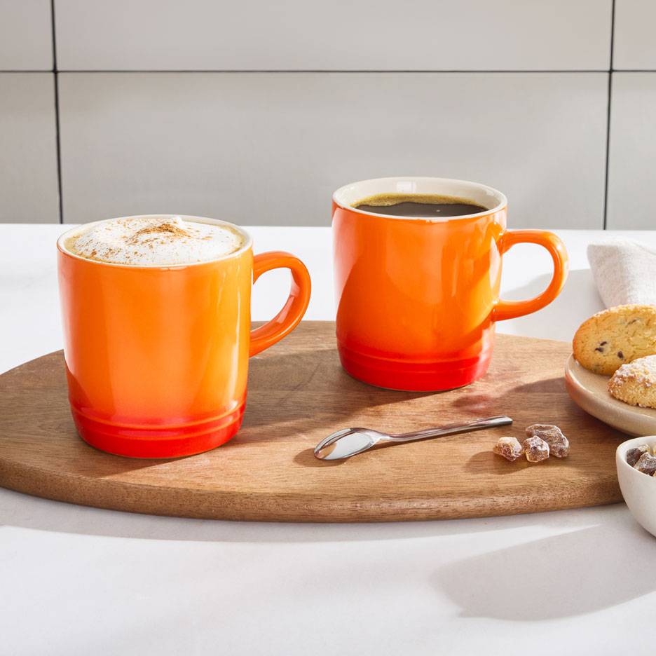 Twee oranje koffiemokken op een houten dienblad, één met schuimige koffie, de andere met zwarte koffie.