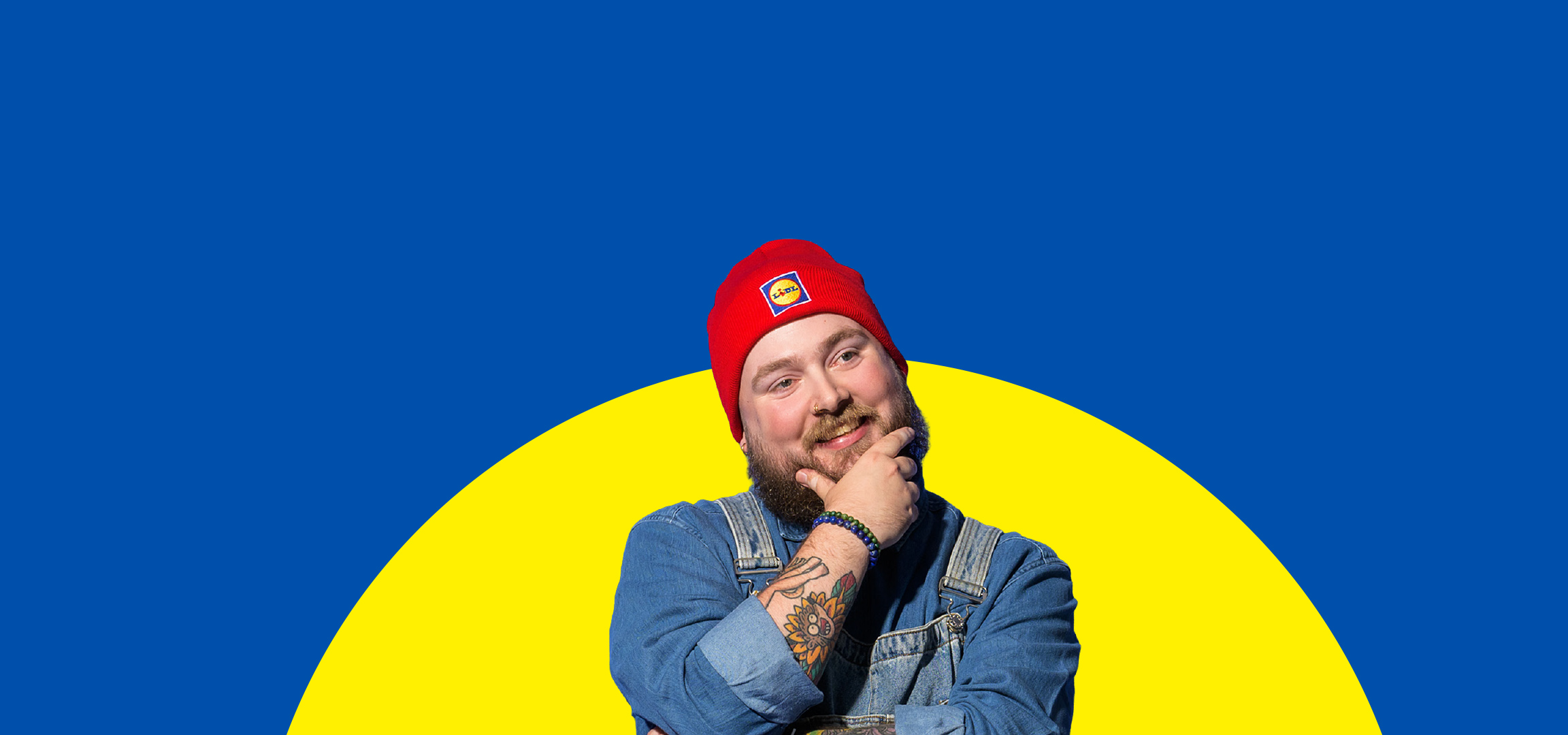 Een lachende man met baard en rode muts met logo, in een spijkeroveral, op een blauw-gele achtergrond.
