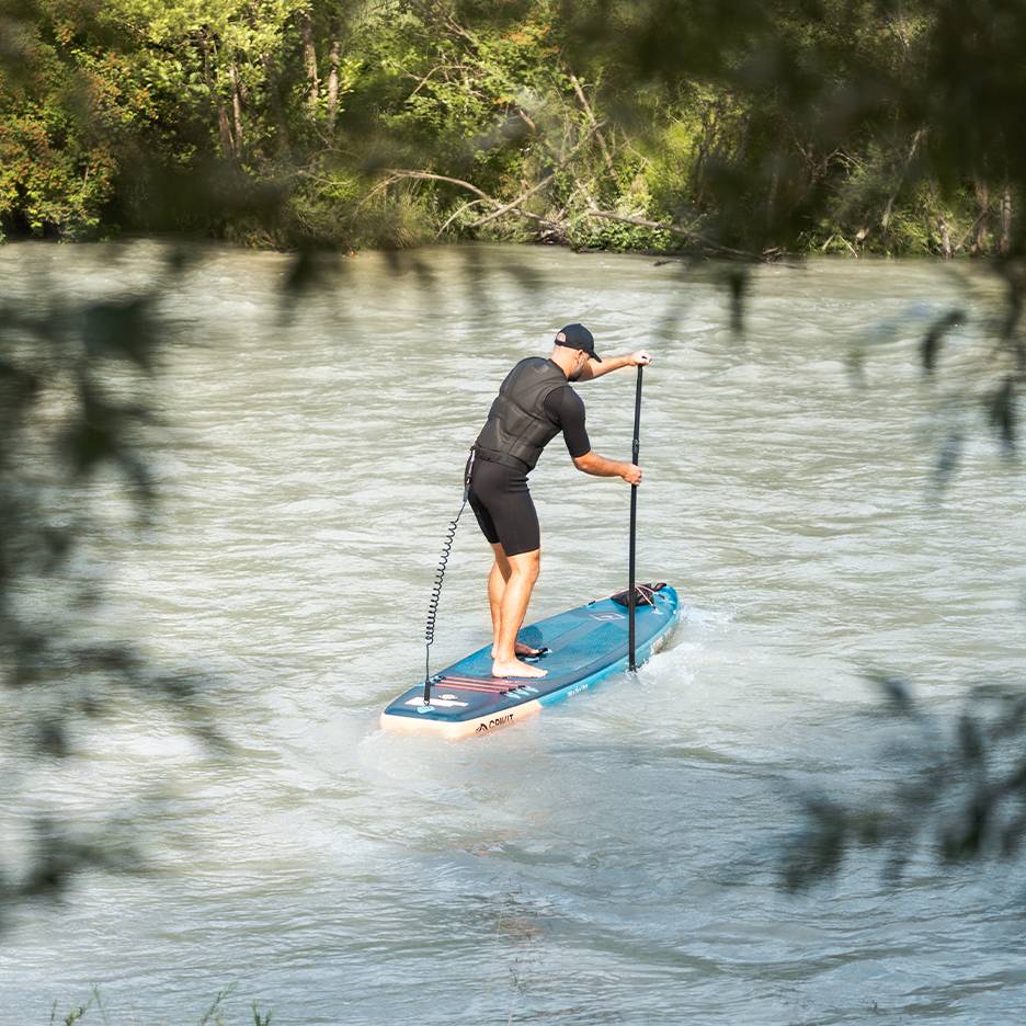 Man in wetsuit en zwemvest peddelt op een blauw paddleboard op een rivier.