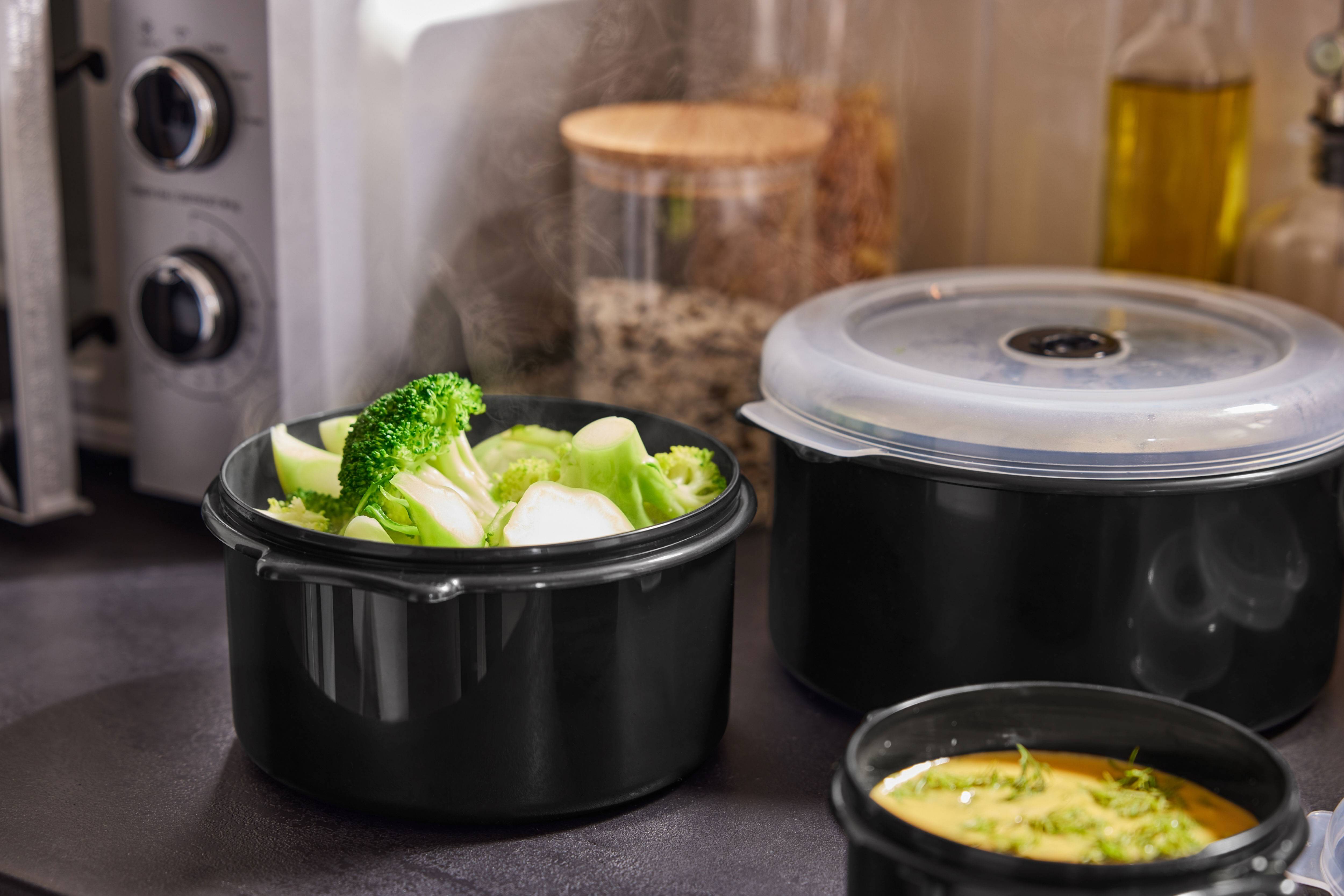 Stoomende broccoli en andere gerechten in zwarte containers op een aanrecht.
