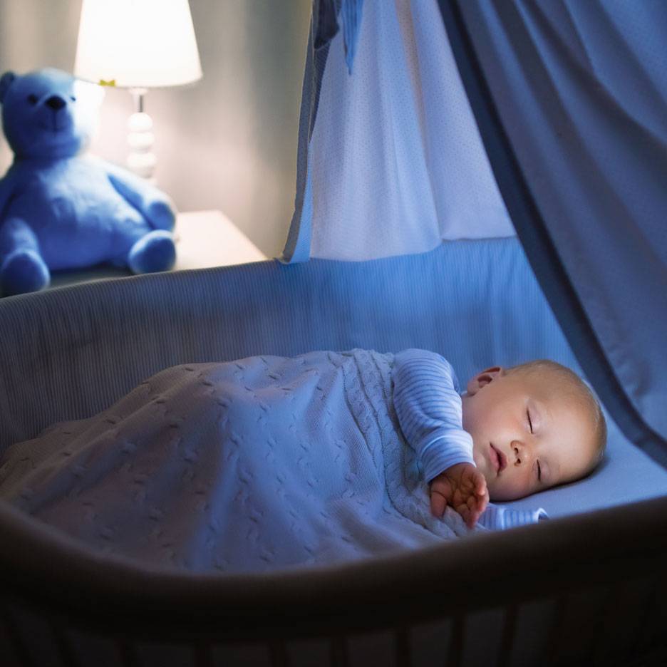 Slapende baby in wieg met blauwe teddybeer en nachtlampje.