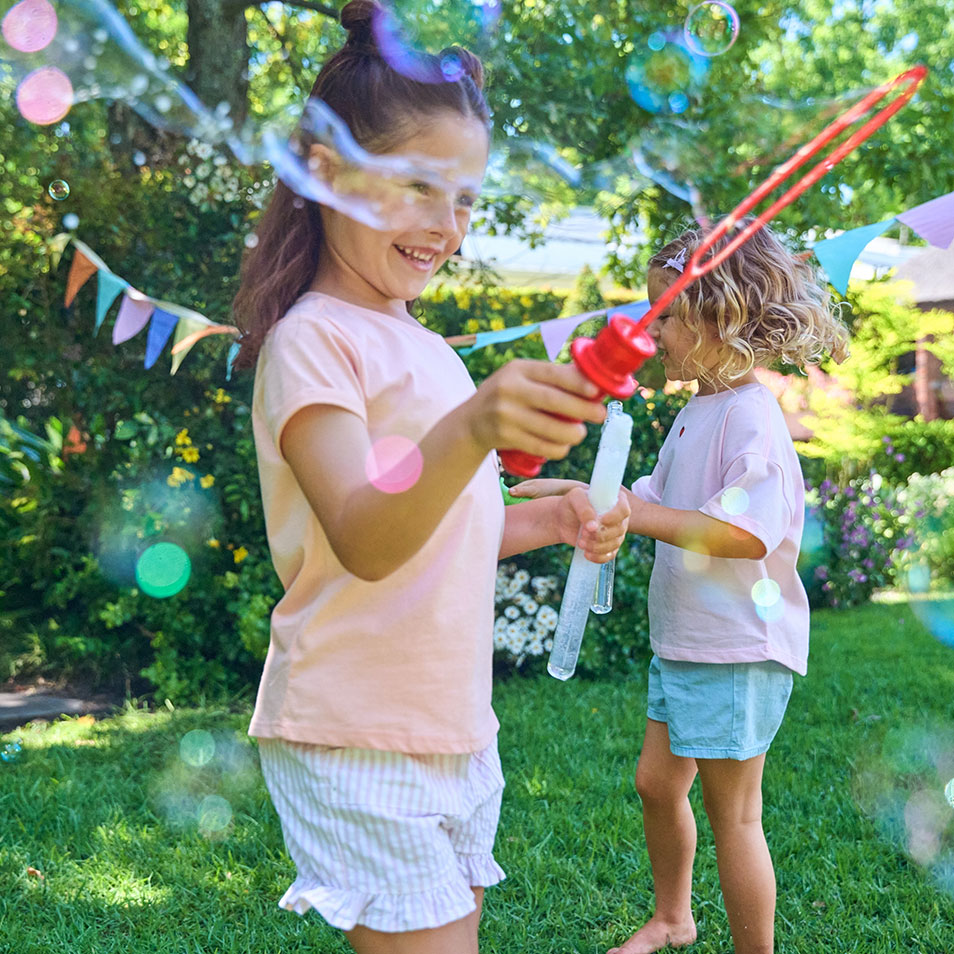 Twee meisjes spelen met bellenblazers in een tuin met kleurrijke vlaggetjes.
