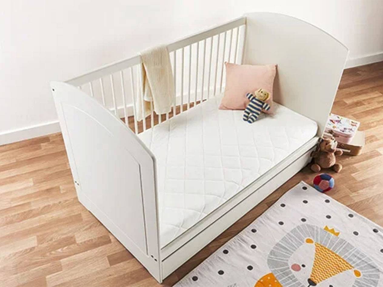 Wit babybedje met matras, kussen en speelgoed op een houten vloer.