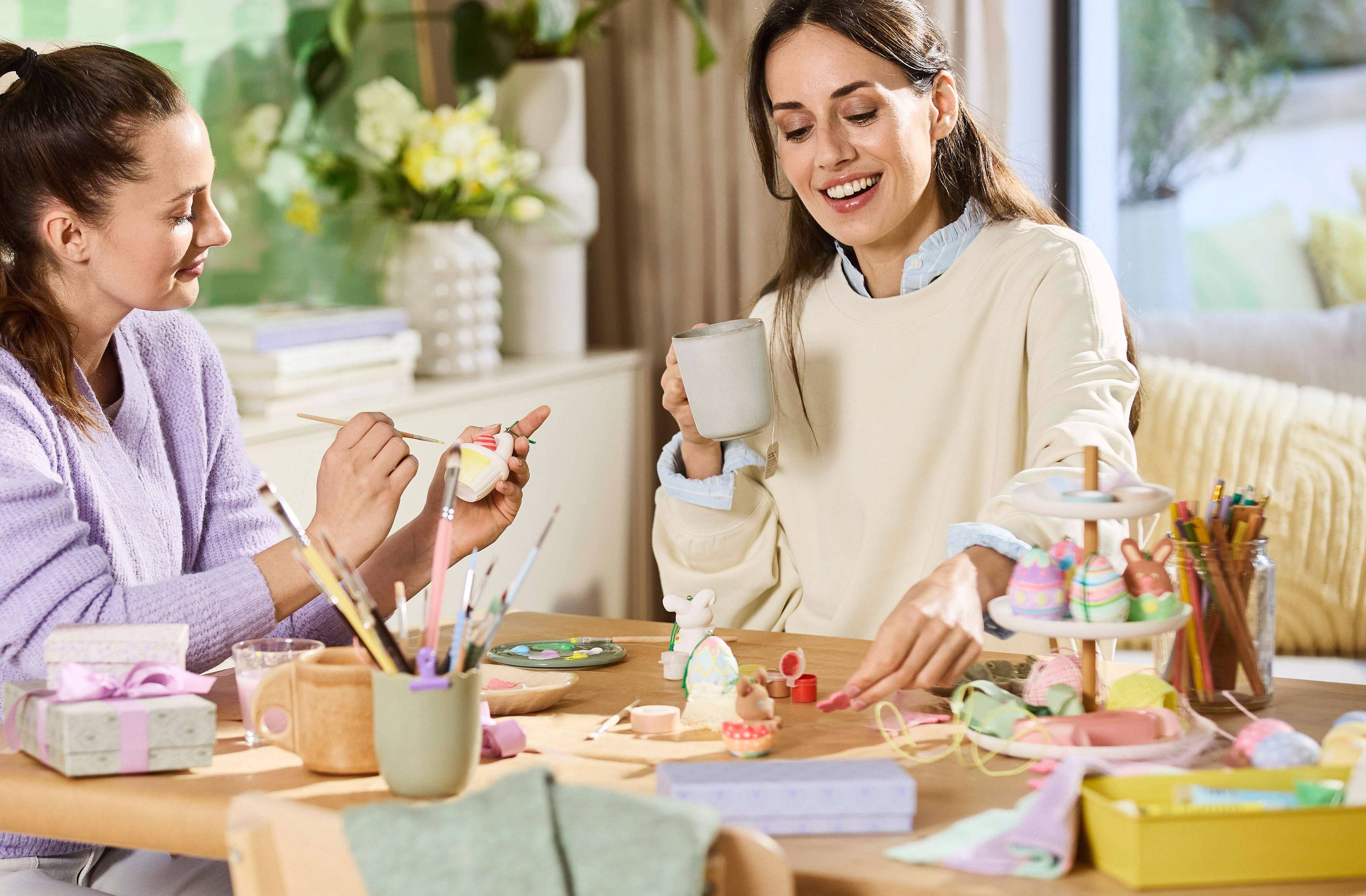 Twee vrouwen beschilderen paaseieren en decoraties, tafel vol knutselspullen.