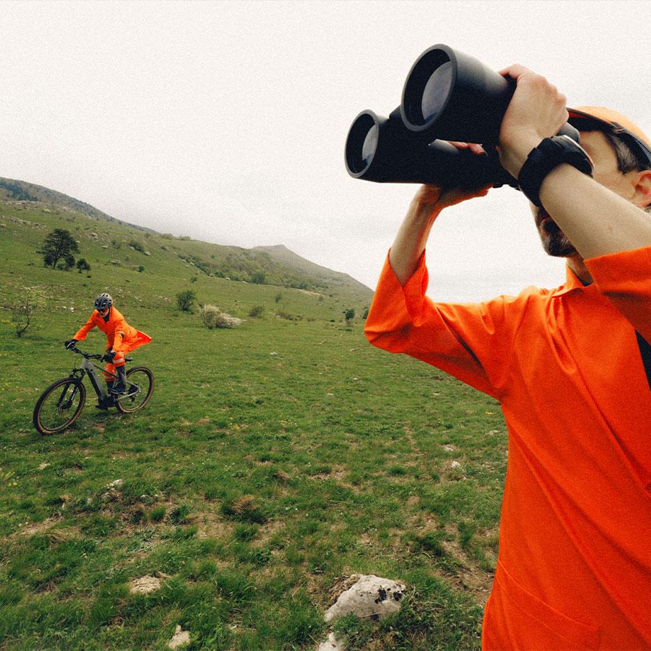 Man met verrekijker en fietser in oranje sportkleding in berglandschap.