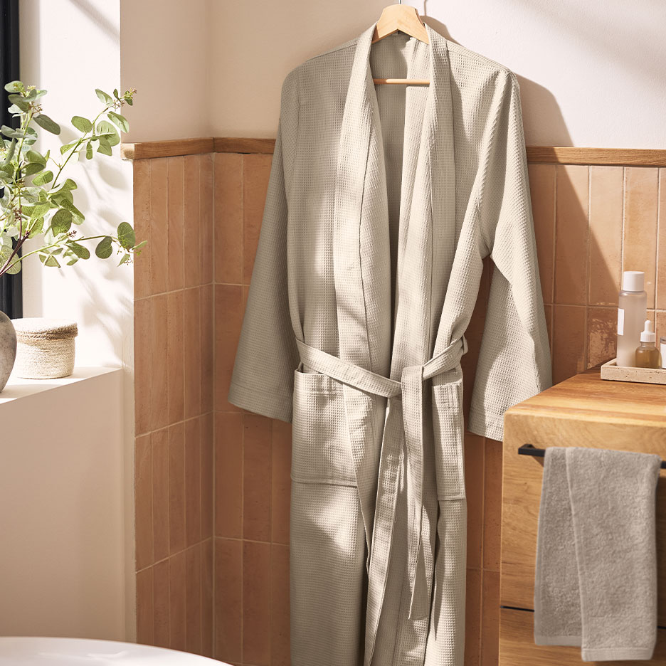 Beige wafelbadjas hangend in een badkamer met bruine tegels en planten.