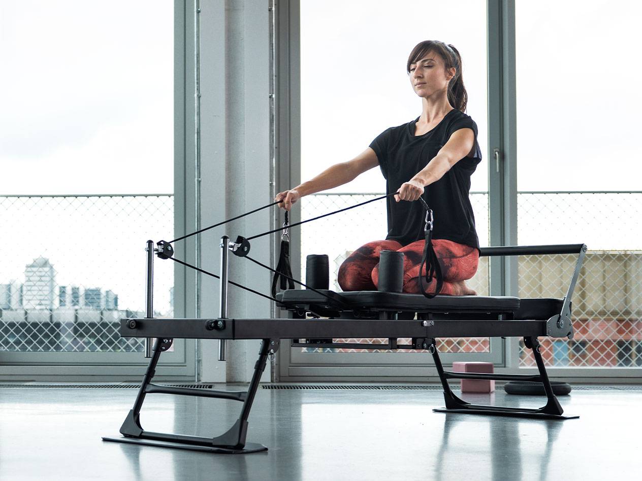 Vrouw doet Pilates op een reformer machine met stadsachtergrond.