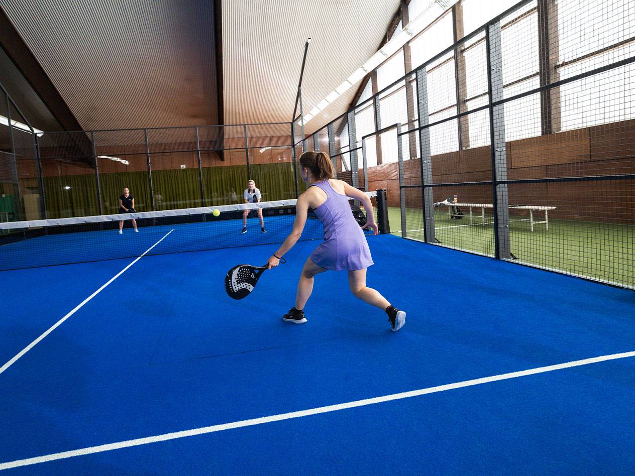Vrouw speelt padel op een blauwe baan, met andere spelers op de achtergrond.