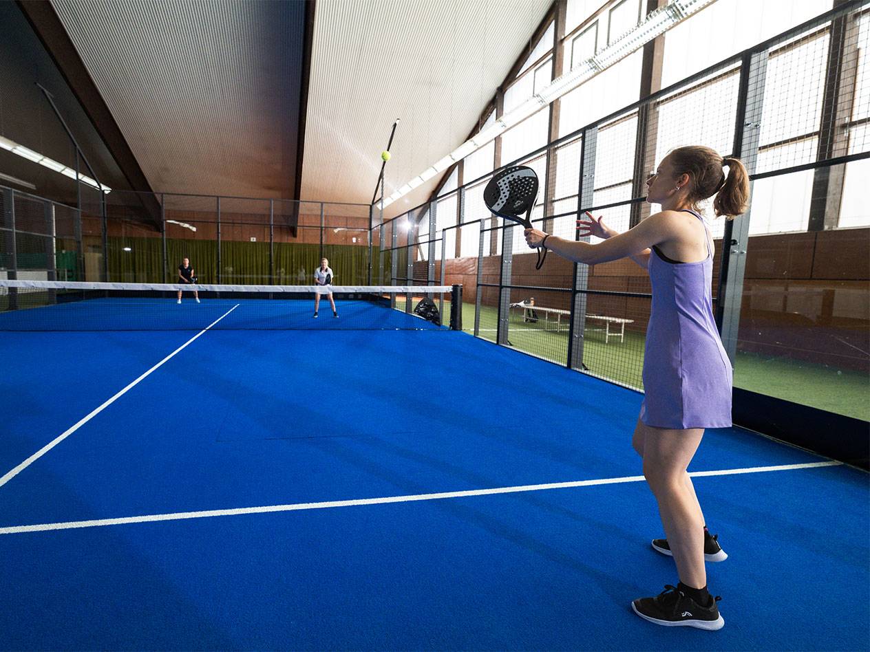 Vrouw in paarse jurk speelt padel op blauw veld, andere spelers op de achtergrond.