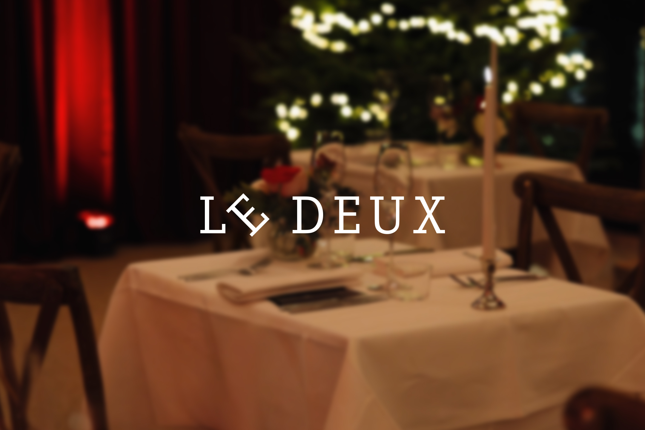 Een restauranttafel met witte kleden en kaarsen, met de tekst 'LE DEUX' in het midden.