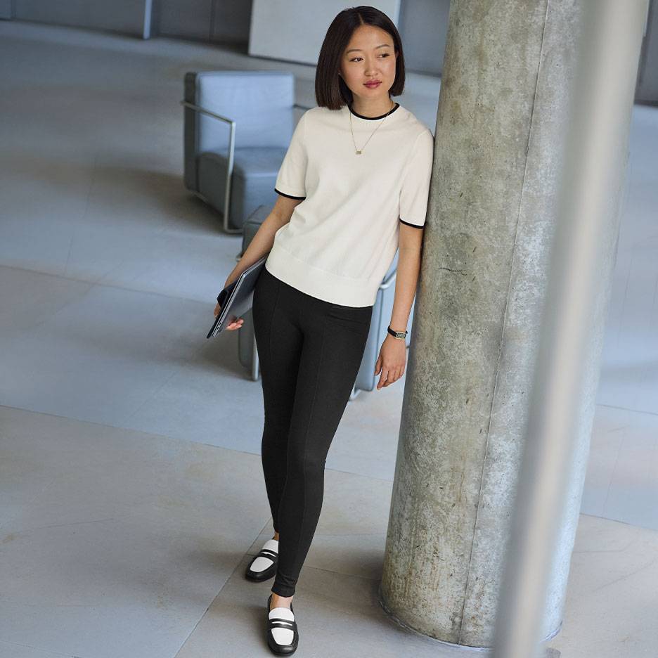 Vrouw in een witte top met korte mouwen en zwarte bies, zwarte legging en tweekleurige loafers.