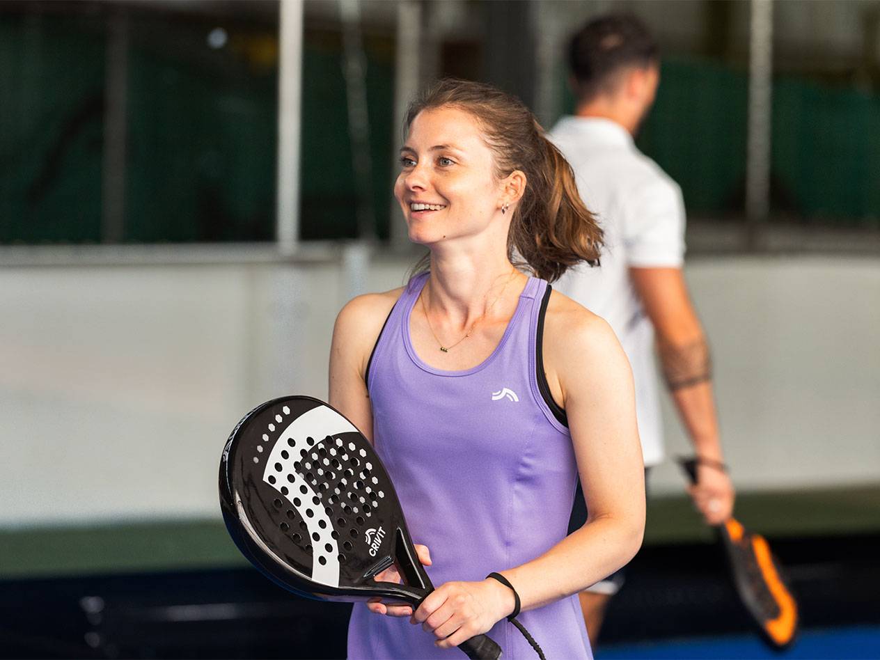 Vrouw in paarse sportjurk met padelracket, man op de achtergrond.