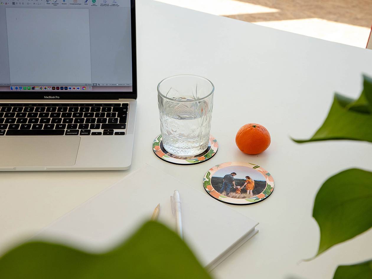 Laptop, glas water, mandarijn en onderzetters met foto's op een wit bureau.