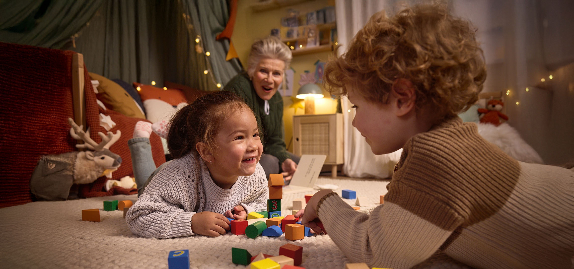 Twee kinderen spelen met kleurrijke bouwblokken op de grond, met een volwassene die toekijkt.