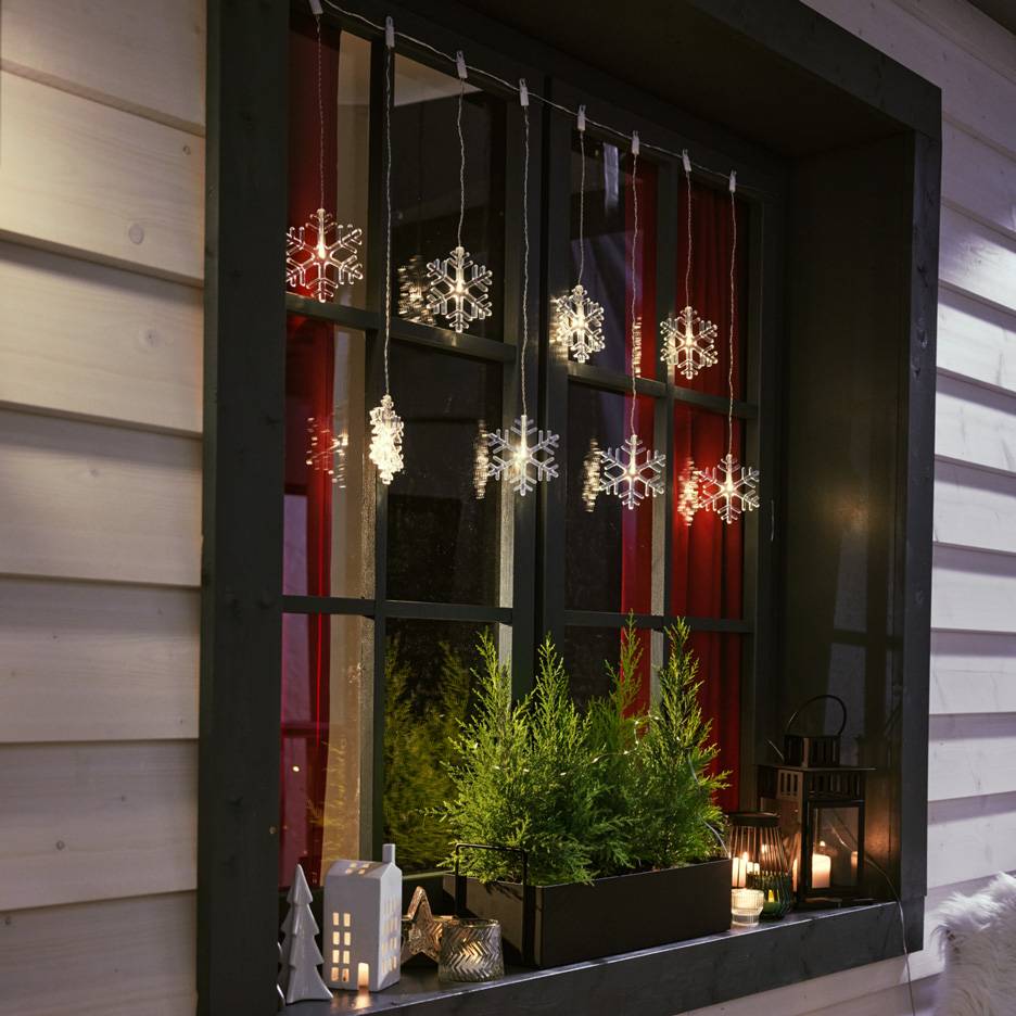 Sneeuwvlok lichtslinger in een raam met kleine dennenboompjes en kaarsen op de vensterbank.