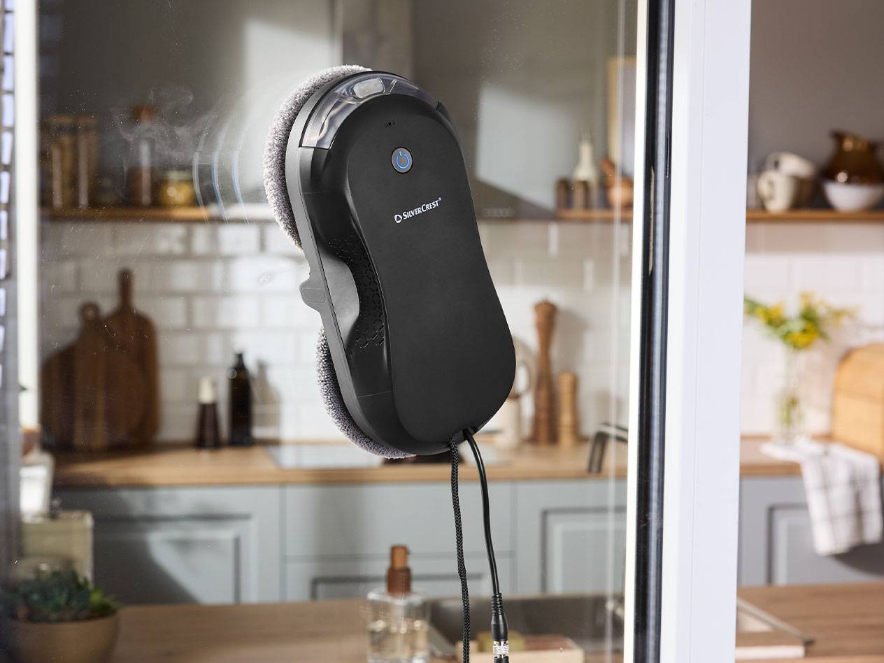 Silvercrest raamreinigingsrobot op een raam met een keuken op de achtergrond.