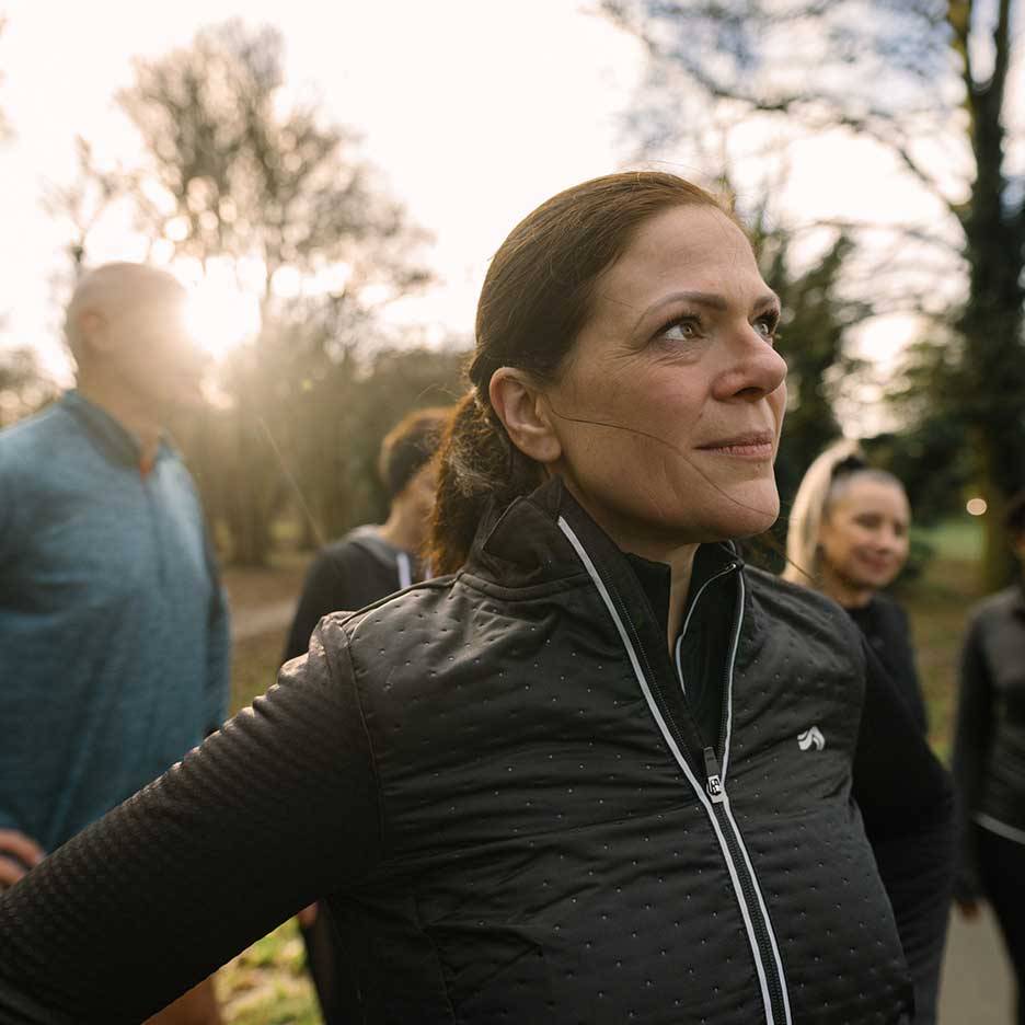 Vrouw in zwart sportjack kijkt omhoog, met een groep mensen op de achtergrond.
