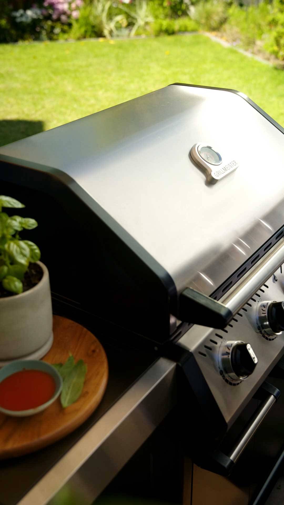Roestvrijstalen gasbarbecue met thermometer, naast een basilicumplant en saus.