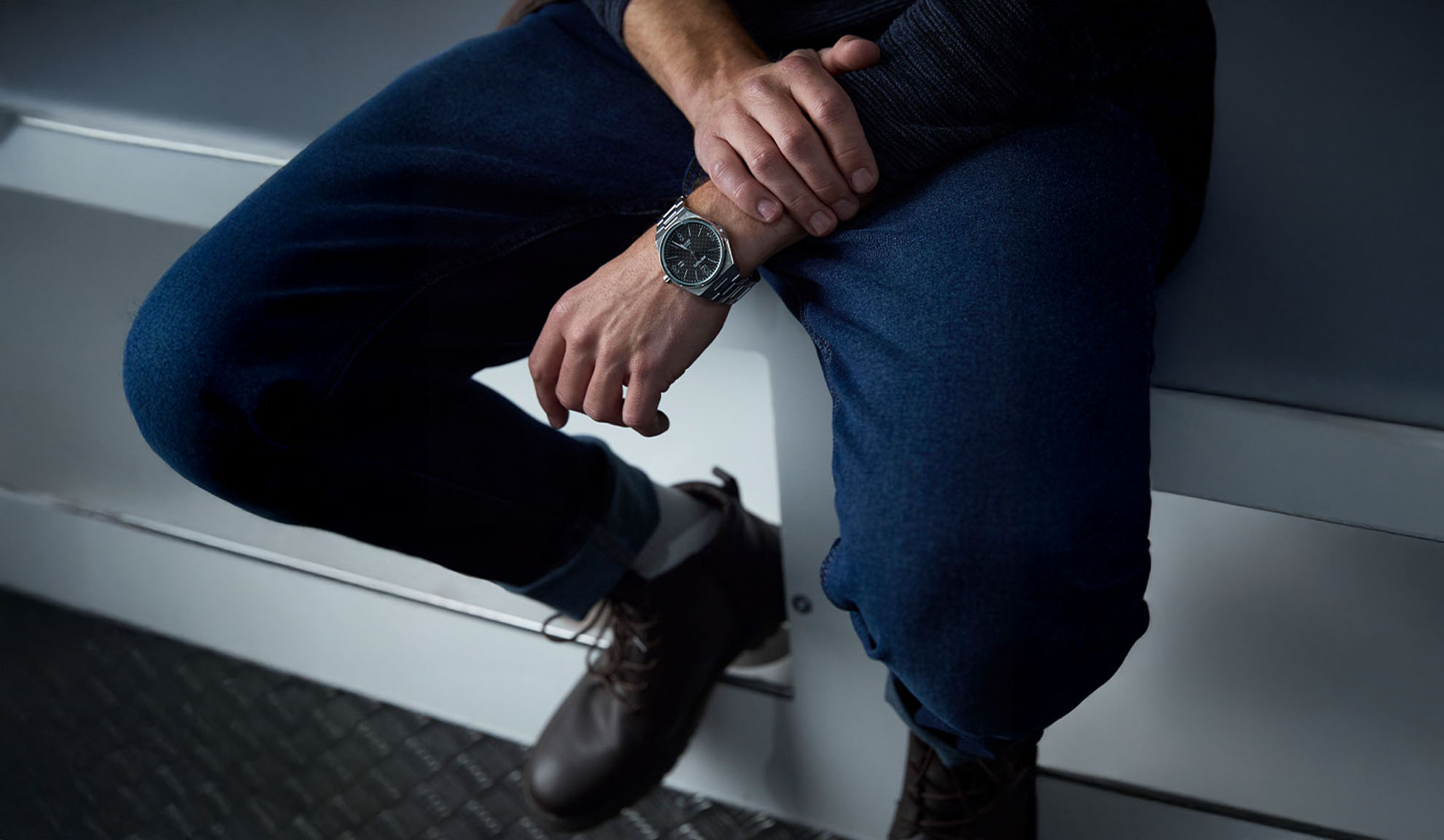 Man draagt donkerblauwe jeans, een donkere trui en een zilveren horloge, zittend.