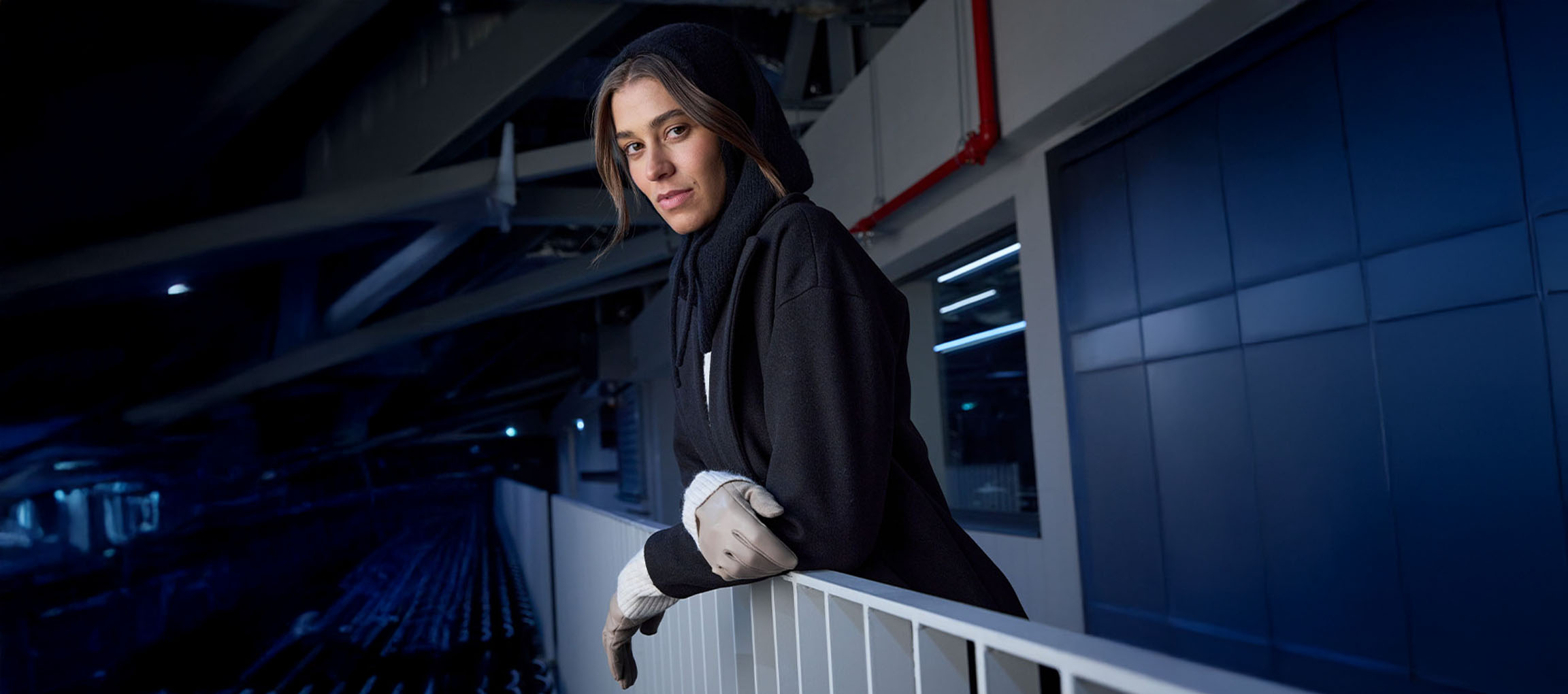 Vrouw in zwarte jas en handschoenen leunend op een reling in een stedelijke omgeving.