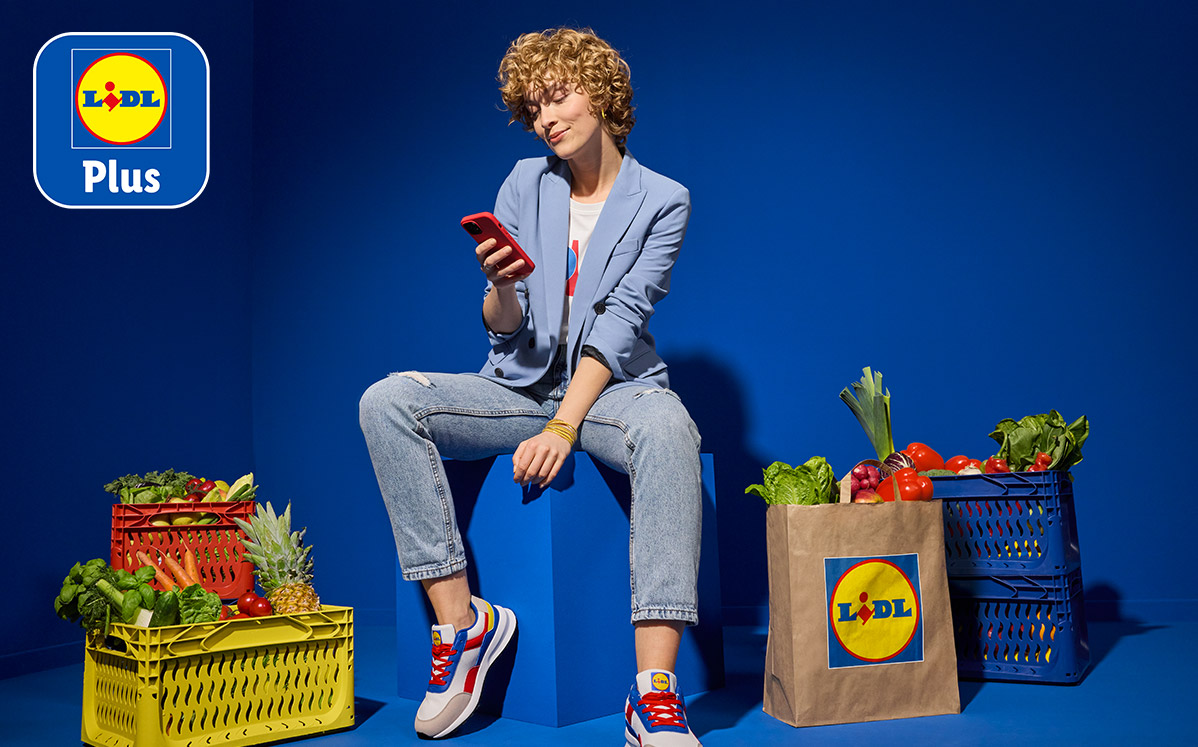 Een vrouw zit op een blauwe kubus en kijkt op haar telefoon, omringd door kratten en tassen vol boodschappen van Lidl.