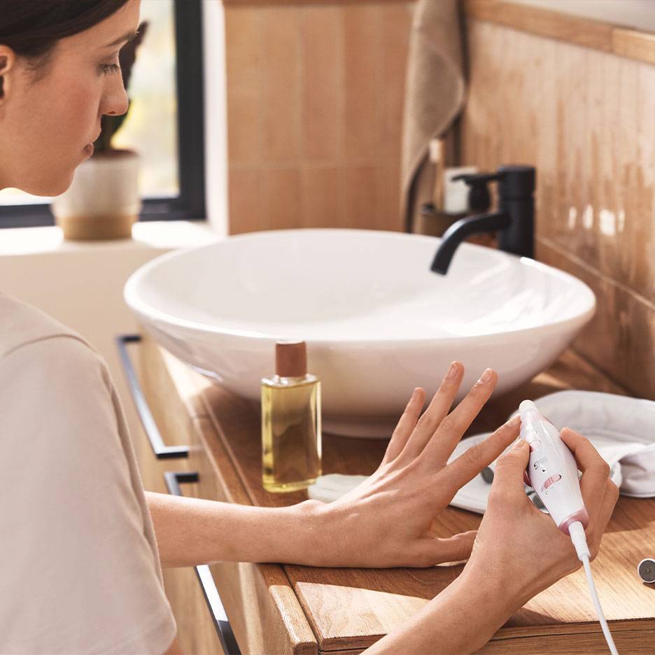 Vrouw gebruikt een elektrische manicure/pedicure set bij een houten wastafelmeubel met witte wasbak.