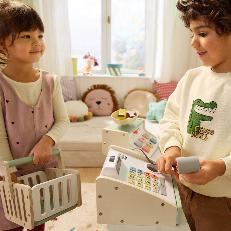 Kinderen spelen winkeltje; jongen bedient kassa, meisje draagt boodschappenmand.