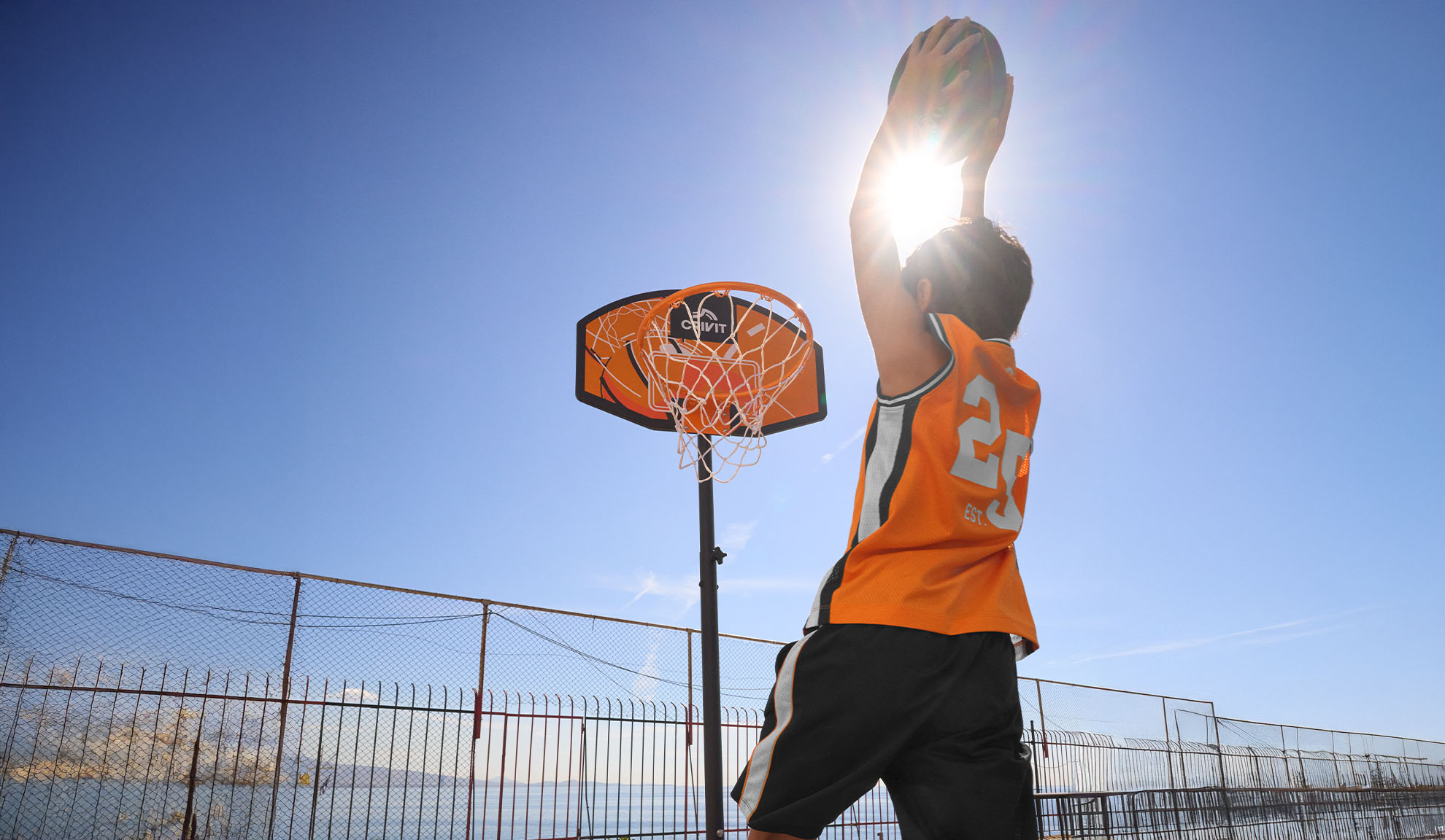 Jongen in basketbalshirt schiet buiten op de basket onder een felle zon.