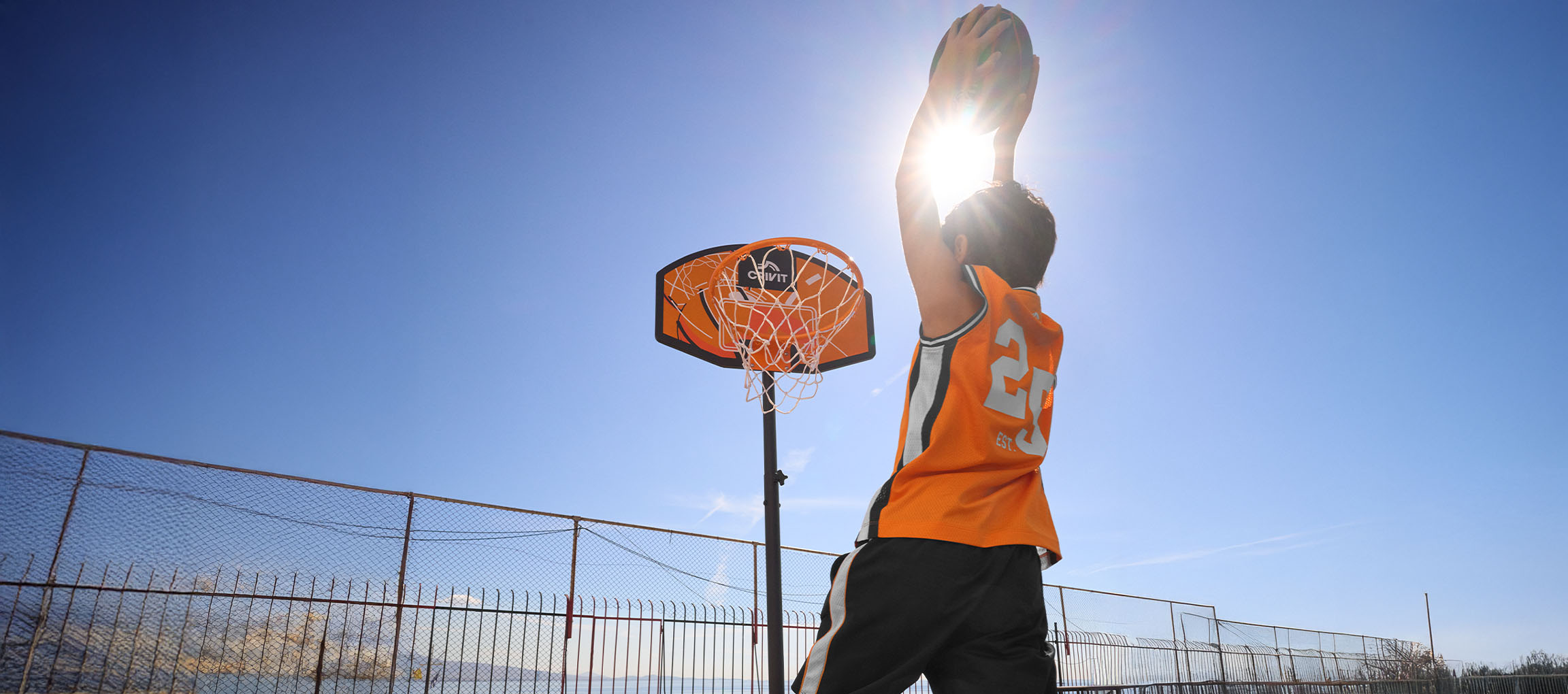 Jongen in oranje shirt gooit bal in basketbalring tegen blauwe lucht.