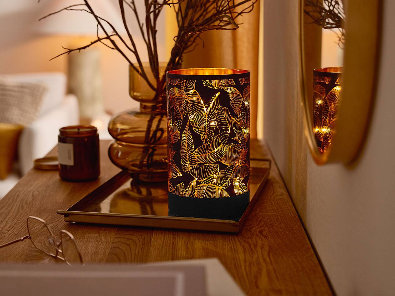 Een decoratieve LED-lantaarn met bladpatroon, een vaas met takken en een kaars op een houten tafel.