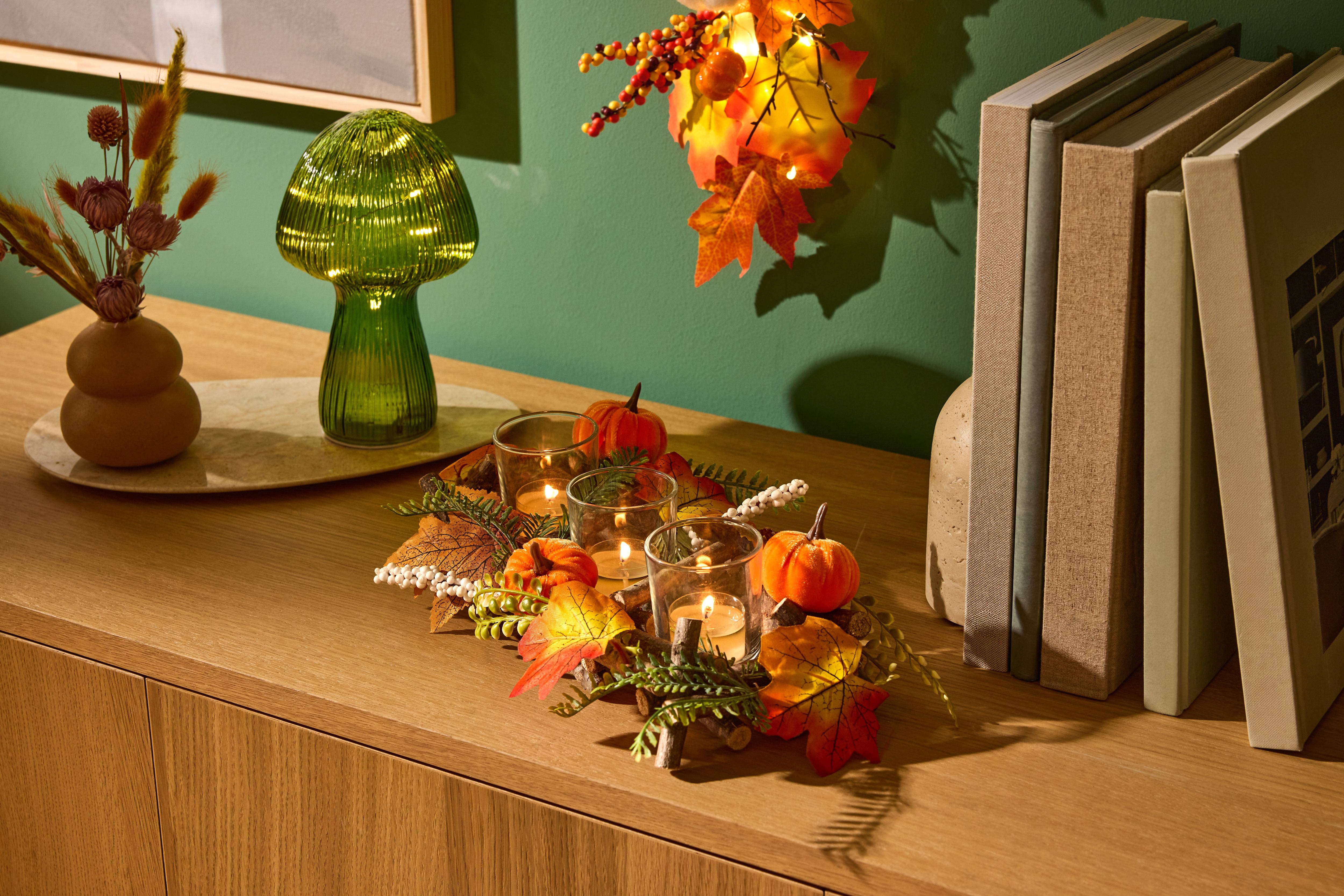 Herfsttafeldecoratie met pompoenen, kaarsen en bladeren, naast een groene paddestoellamp en boeken.