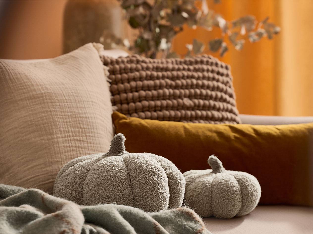 Twee pluche decoratieve pompoenen en kussens in aardetinten op een bank.