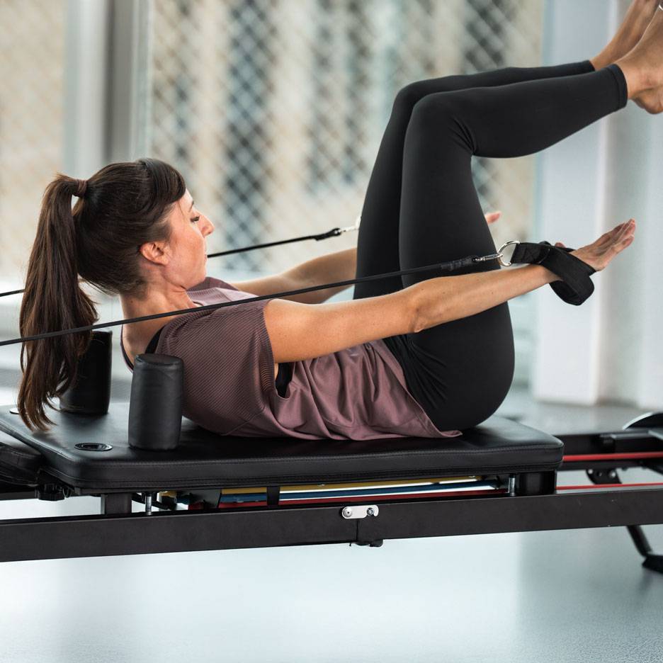Vrouw doet Pilates op een reformer, gekleed in zwarte sportkleding.