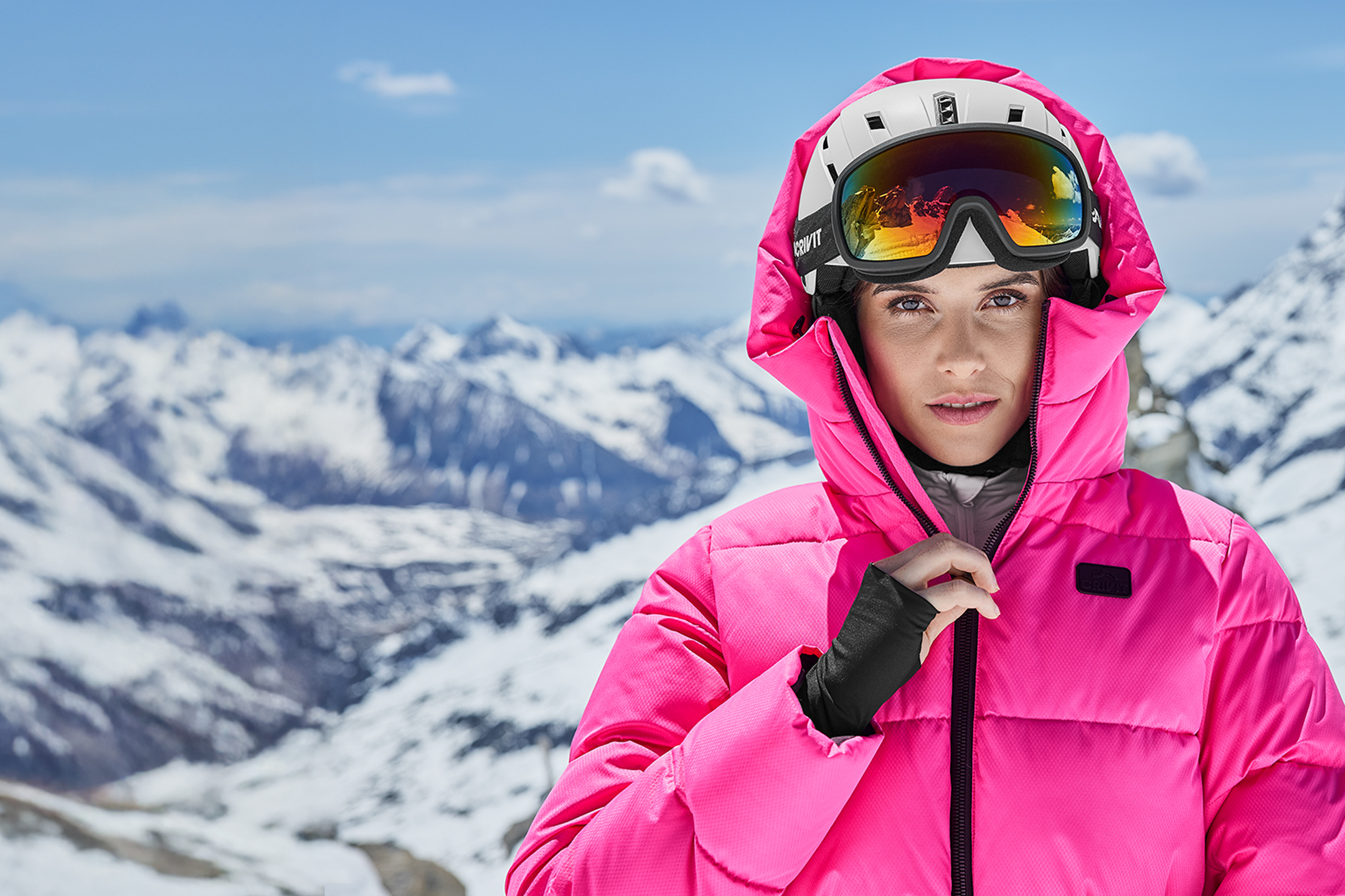 Vrouw in roze ski-jas en skihelm met skibril voor een besneeuwd berglandschap.