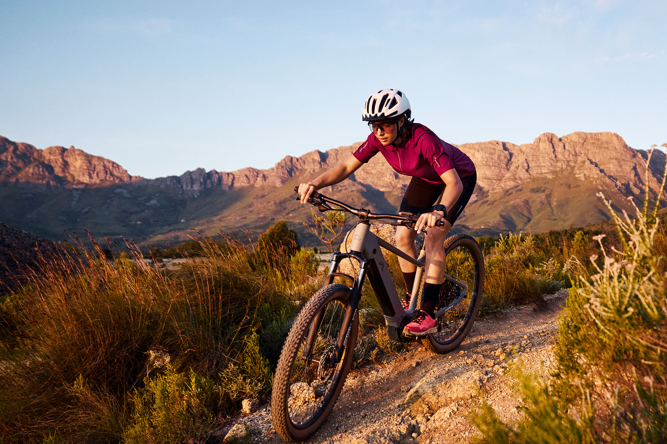 Vrouw mountainbiket op een onverhard pad met bergen op de achtergrond.
