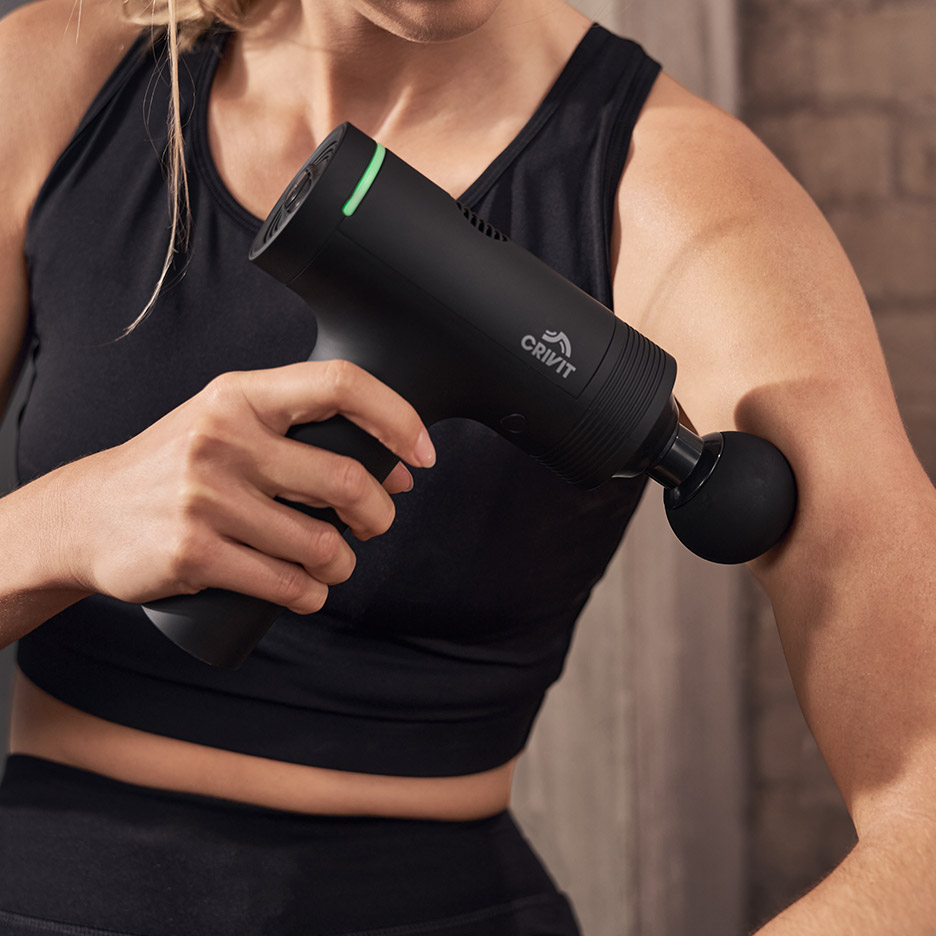 Vrouw in sportkleding gebruikt een zwart CRIVIT massageapparaat op haar bovenarm.