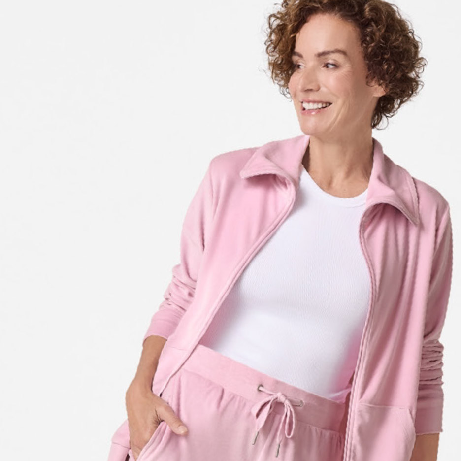 Vrouw in roze trainingspak met ritsvest en broek, lachend en wegkijkend.