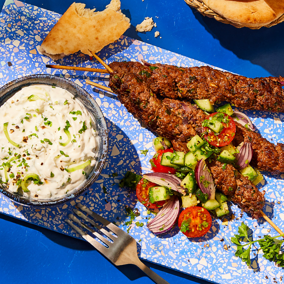 Kebabspiesjes met tzatziki, salade en platbrood op een blauwe ondergrond.