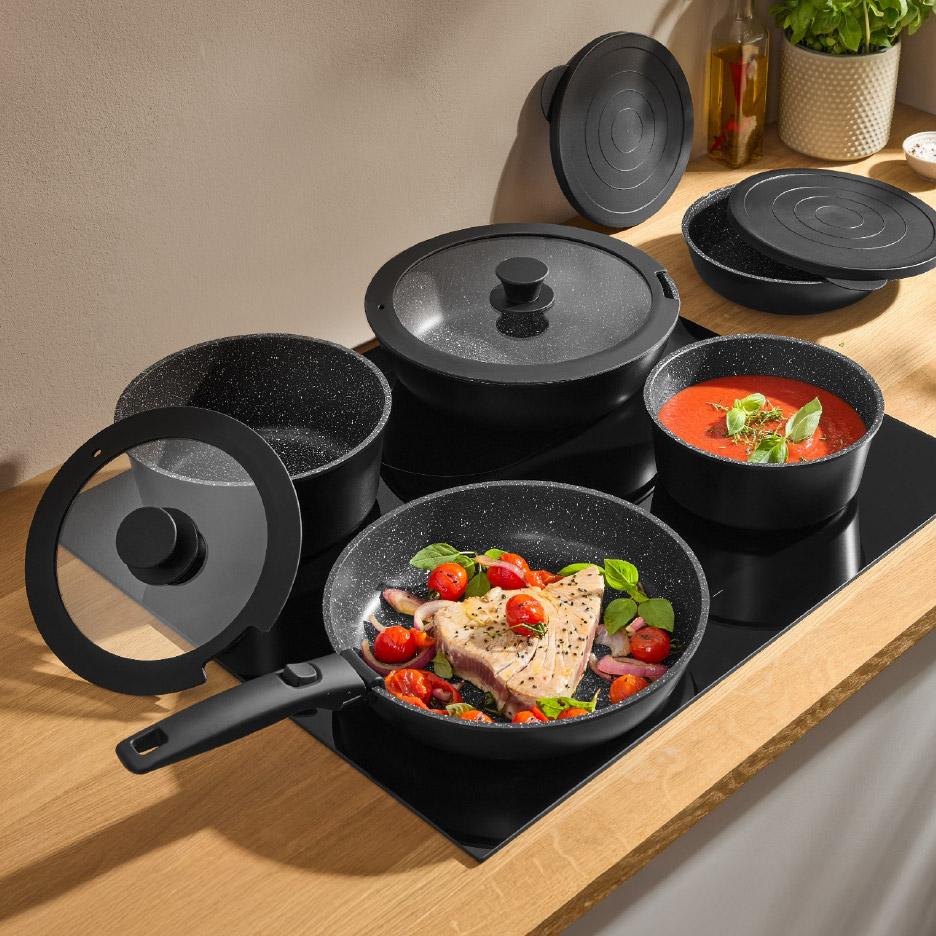 Zwarte pannenset met deksels, inclusief een koekenpan met vis en groenten, en een pan met tomatensaus.