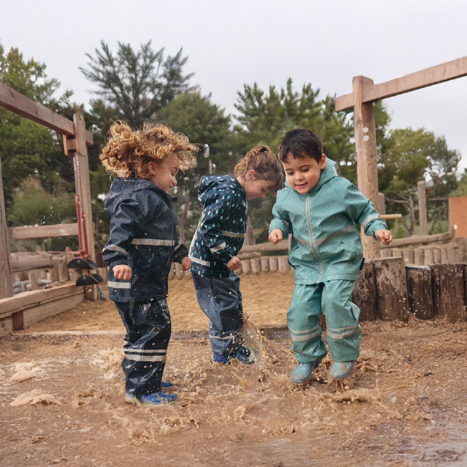 Drie kinderen in regenpakken en regenlaarzen springen in een modderpoel.