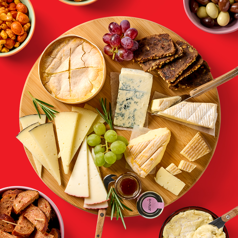 Een houten kaasplank met diverse kazen, druiven, crackers en rozemarijn, omringd door snacks.