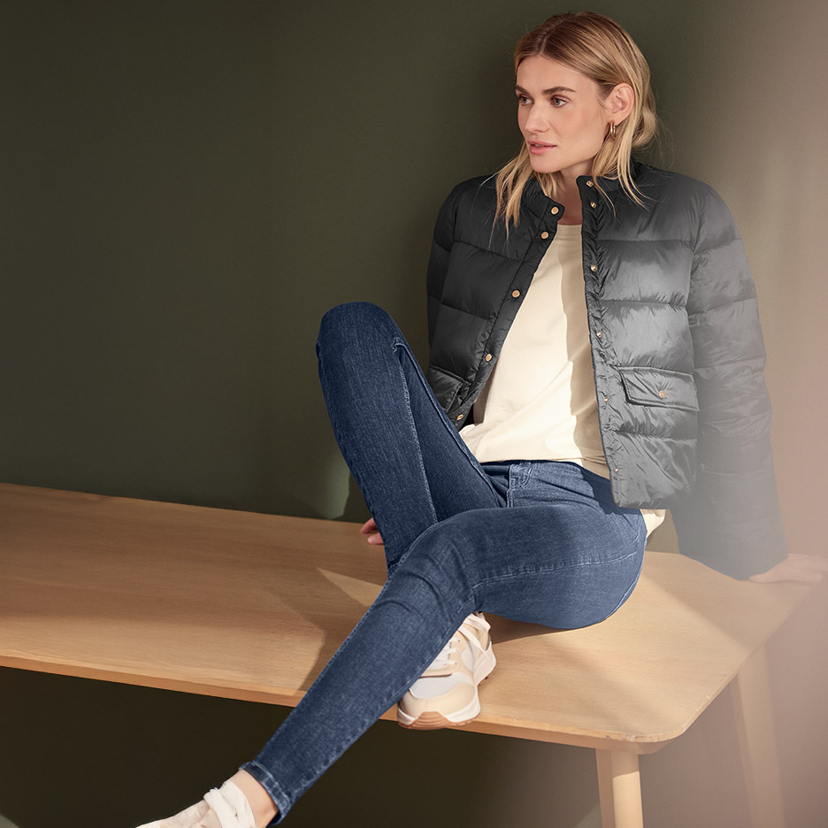 Vrouw in een zwart gewatteerd jack, crème T-shirt en donkerblauwe jeans, zittend op een bank.