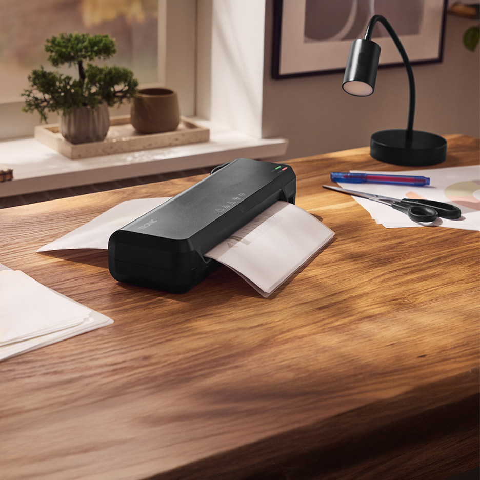 Zwarte lamineermachine op een houten bureau met papier, schaar en een lamp.