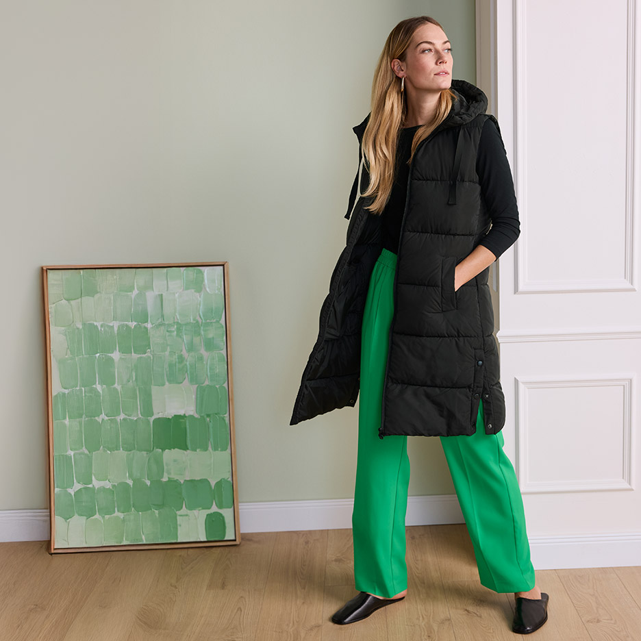 Vrouw in een lange zwarte gewatteerde bodywarmer en felgroene broek, staand naast een schilderij.